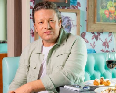 Jamie Oliver sitting off the side of a restaurant booth seat, looking at the camera
