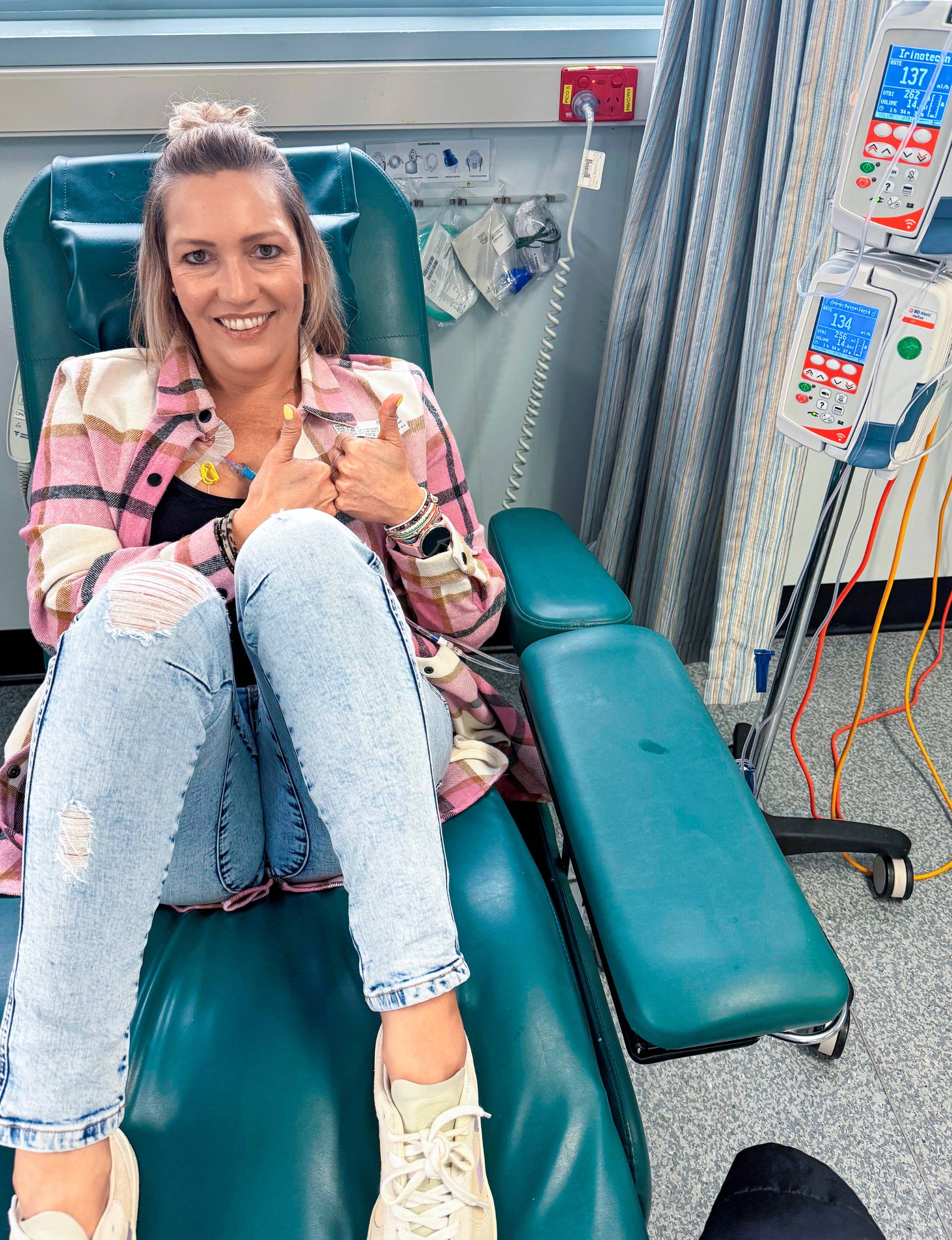 Becki laying on a chemo bed holding two thumbs up