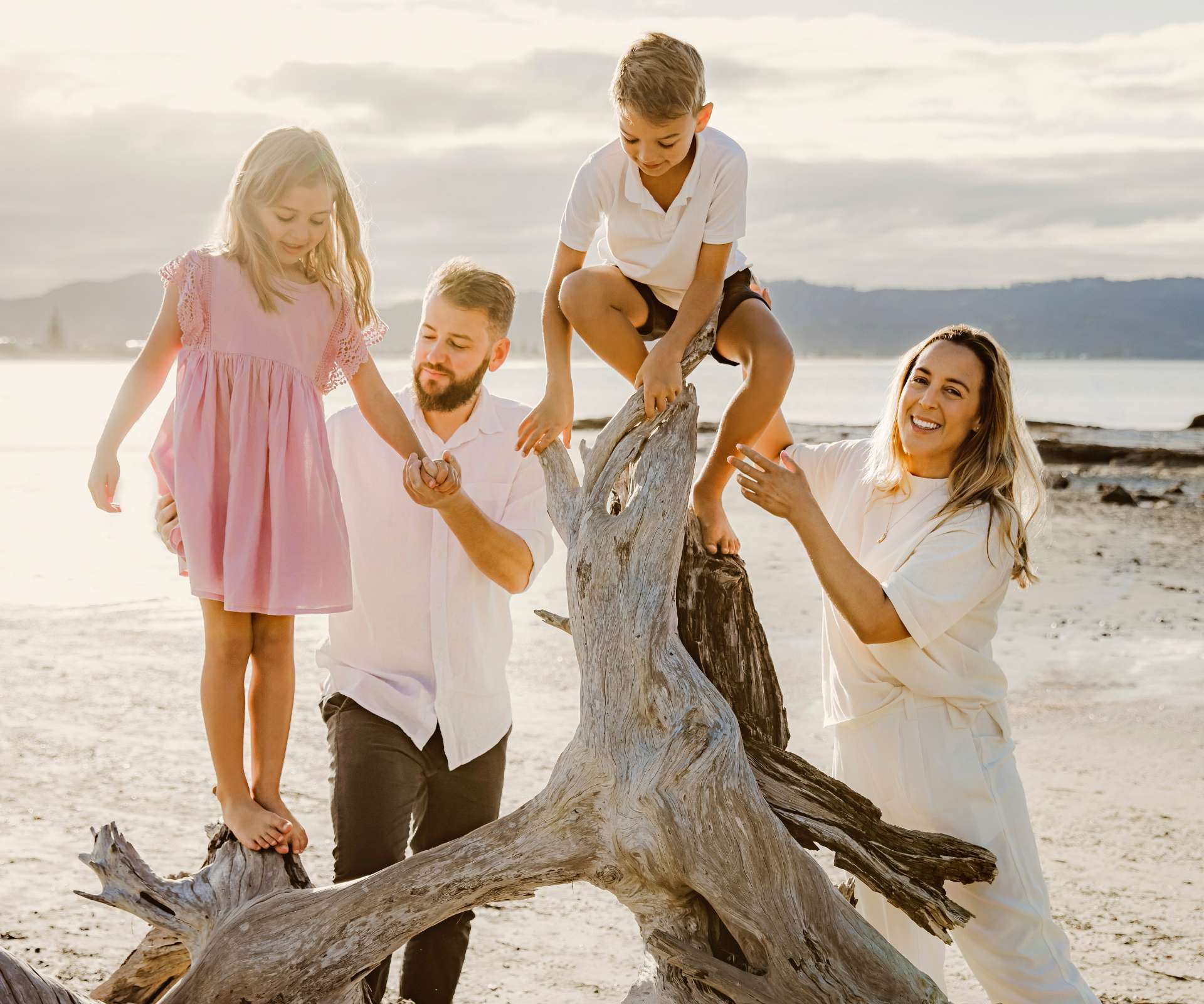 A family photo of Emma and Richard holding their kids while they climb on some driftwood at the beach