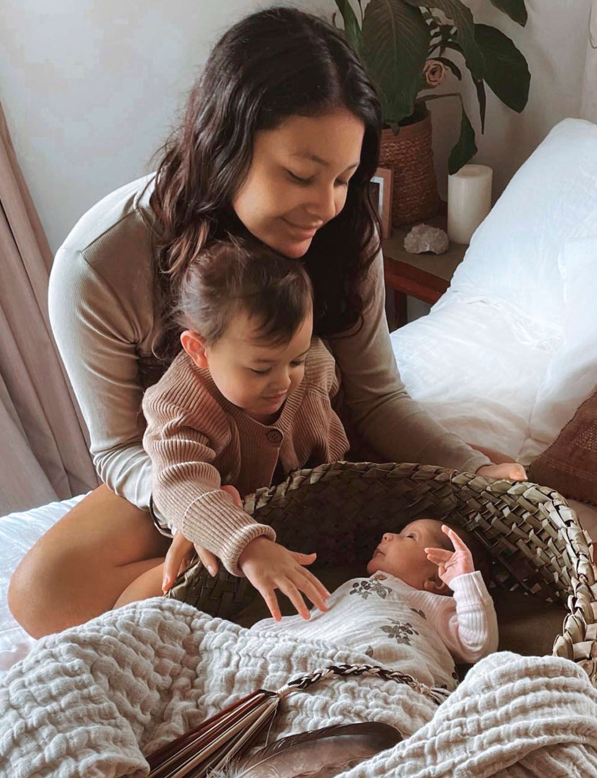 Wikitoria sitting on her bed with with one daughter sitting between her legs, and the youngest in a flax-woven crib in front of them
