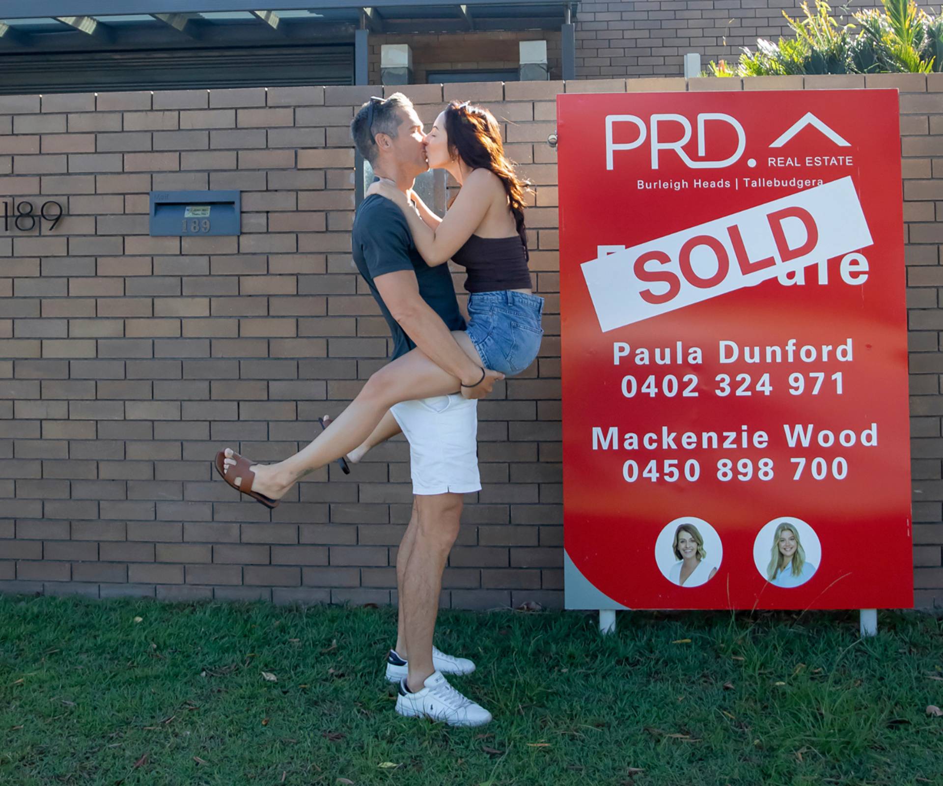 Jono and Ellie kissing in front of the SOLD sign in front if their new home