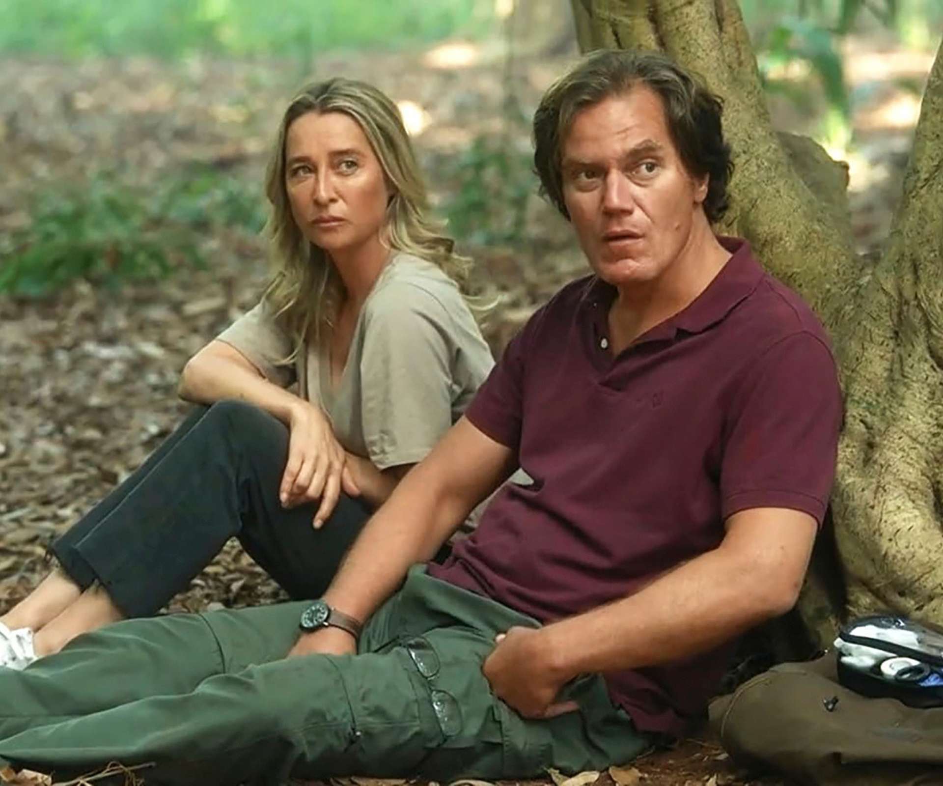 Man and woman sitting on forest floor against a tree