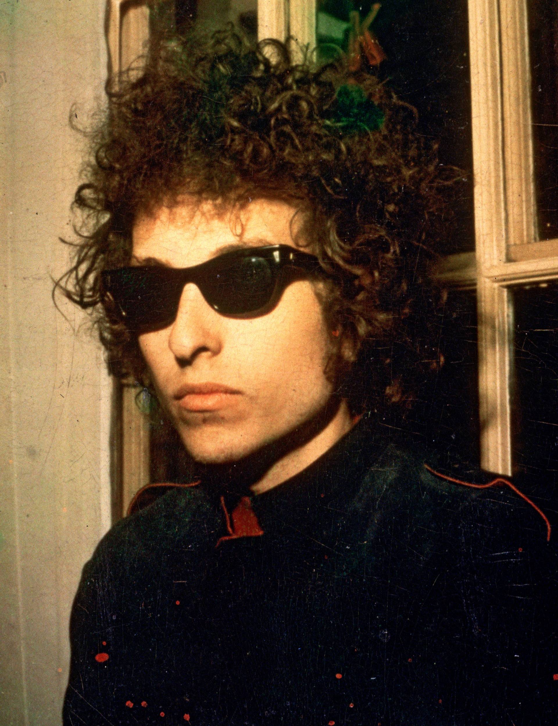 Bob Dylan leaning against a window with sunglasses on at night in 1966