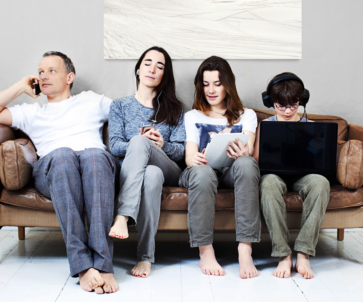 Family of four sit on couch all using electronic devices